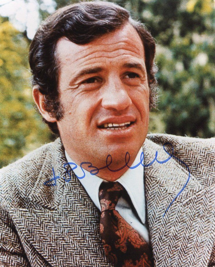 Jean-Paul Belmondo smiling while looking afar and wearing a coat, white long sleeves, and brown necktie