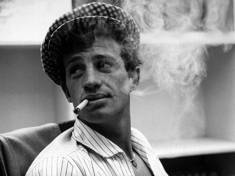 Jean-Paul Belmondo smoking while looking at the back and wearing a checkered cap and striped long sleeves