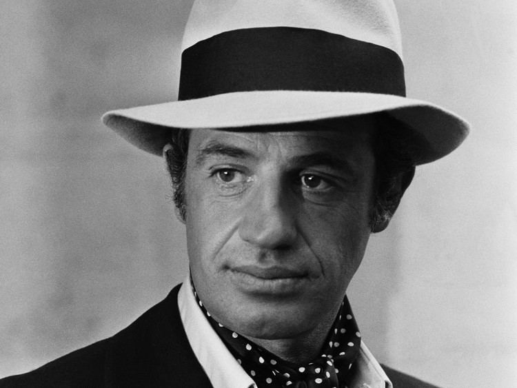 Jean-Paul Belmondo looking afar while wearing a hat, coat, long sleeves, and polka dot ribbon scarf
