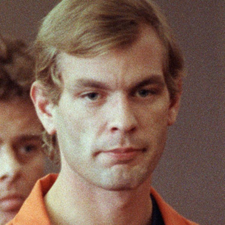 At the back a man is serious, has brown hair, in front, Jeffrey Dahmer is serious, looking down, has blond hair wearing an orange prison uniform.