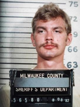 In a police Mug shot with a black lines with numbers at the back to determine the height at the back, in front Jeffrey Dahmer is serious, has brown hair and a mustache, wearing a white shirt, with a black placard with a written “MILWAUKEE COUNTY” “ SHERIFF'S DEPARTMENT” “56580 80882”