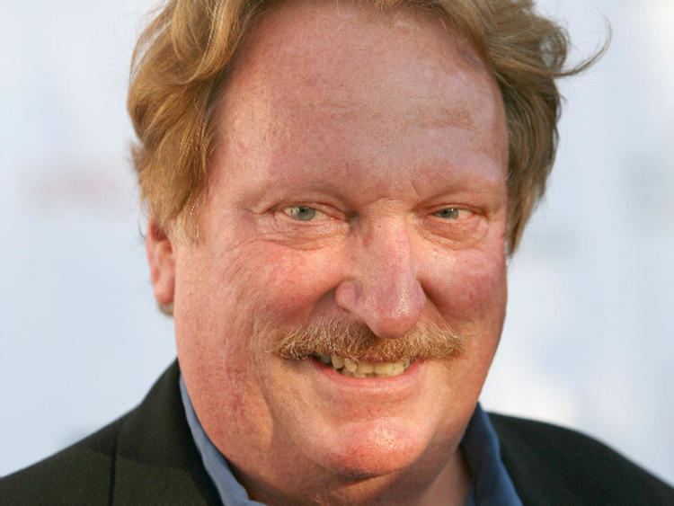 Jeffrey Jones smiles in the white background while wearing a black suit