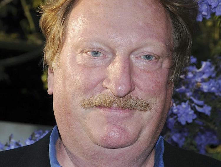 Jeffrey Jones smiles while wearing a black suit with a flower at his back