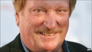 Jeffrey Jones smiles in the white background while wearing a black suit