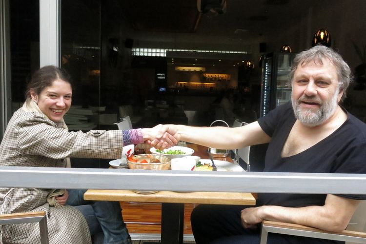 Jela Krečič and Slavoj Žižek are smiling while doing shake hands in a restaurant. Jela wearing a brown coat and blue jeans while Slavoj wearing a black shirt and black pants.