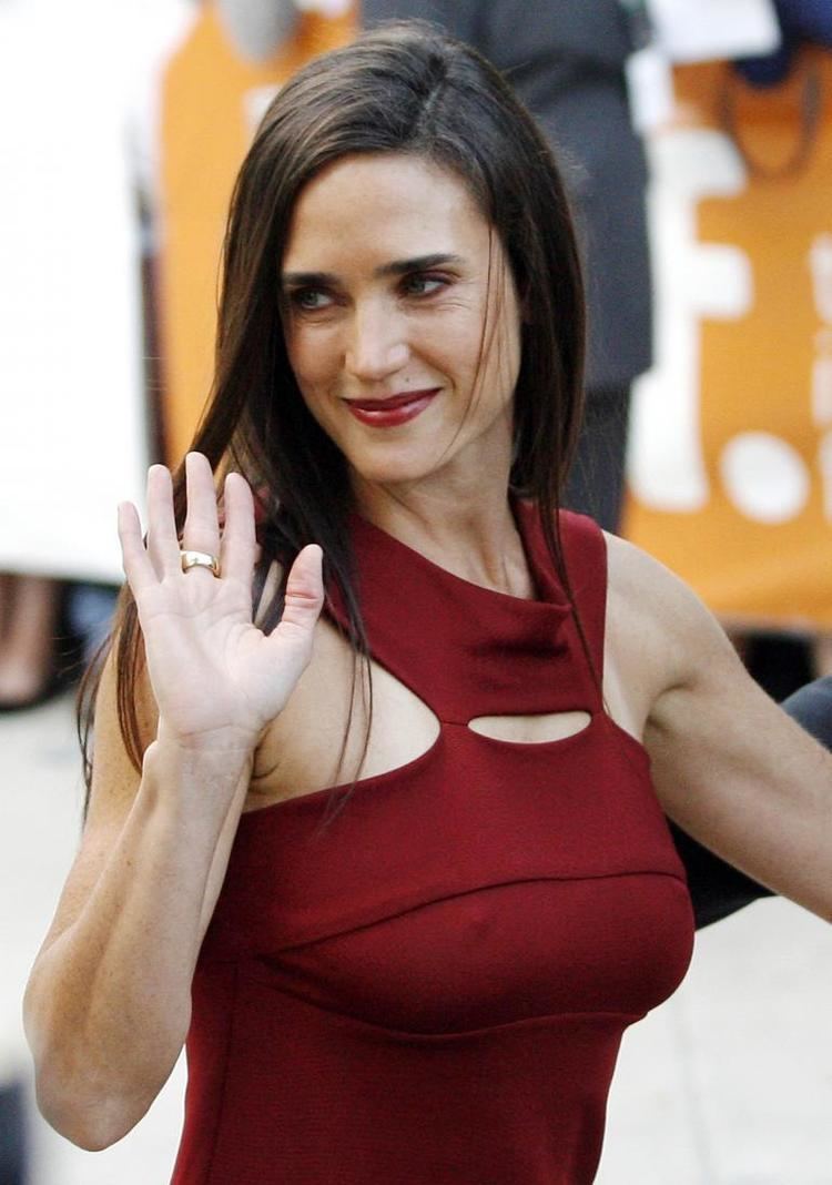 Jennifer Connelly is smiling and waving with her right hand, has long brown hair and mole on her left upper lips, wearing a Gold ring and red dress.