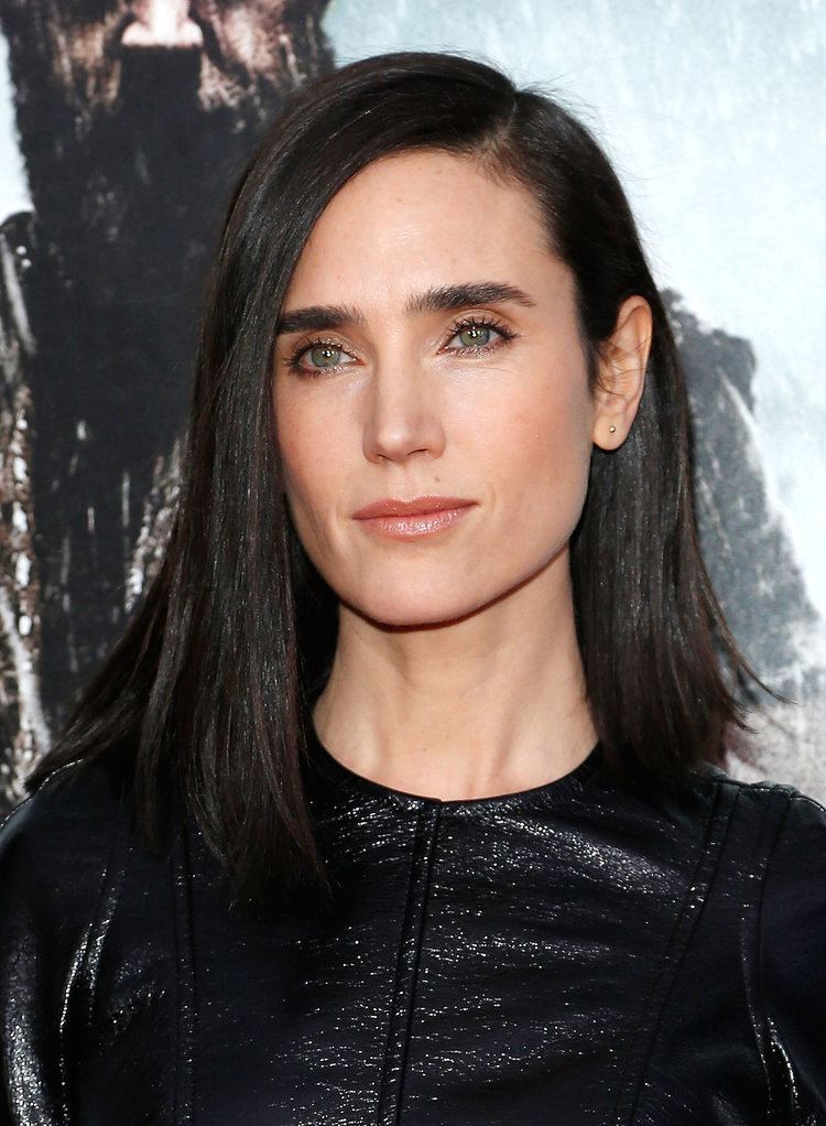 Jennifer Connelly is serious, has long black hair with side bangs, brown eyes and mole on her left upper lips wearing a shiny black top and gold stud earrings.