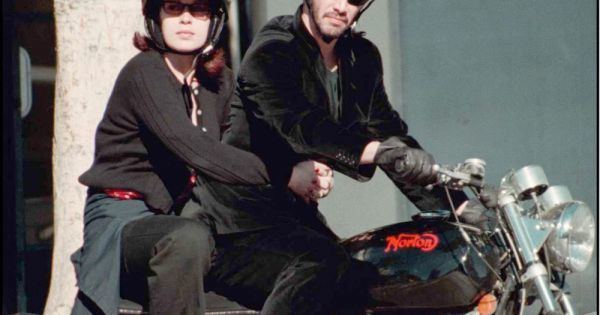 Jennifer Syme riding a motorcycle with Keanu Reeves while wearing a helmet, shades, black jacket, and black pants