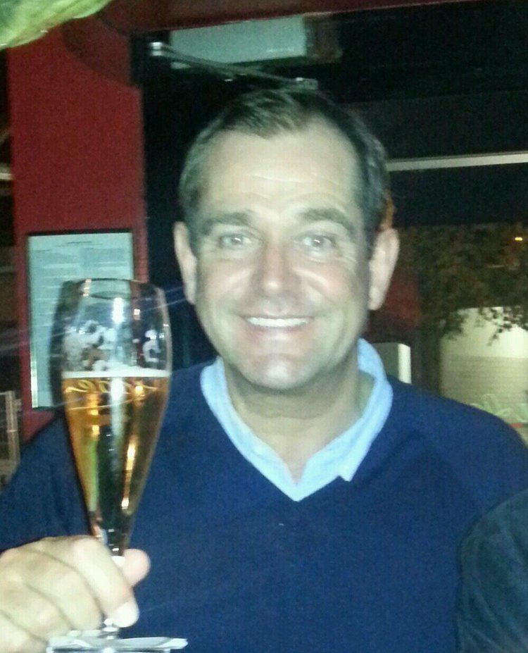 Jeremy Gittins smiling while holding a glass of wine and wearing a dark blue long sleeve shirt and blue polo