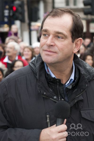 Jeremy Gittins looking afar while holding a microphone and wearing a black jacket and blue polo underneath