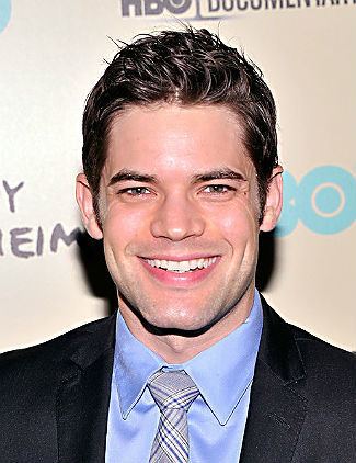 Jeremy Jordan smiling while wearing a black coat, blue long sleeves and necktie
