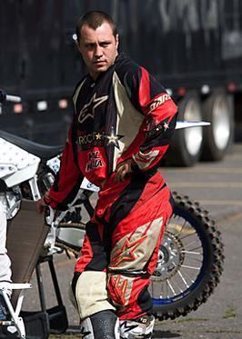 Jeremy in a fierce look standing near a moto racing bike & wearing a red & black colored moto racing dress.