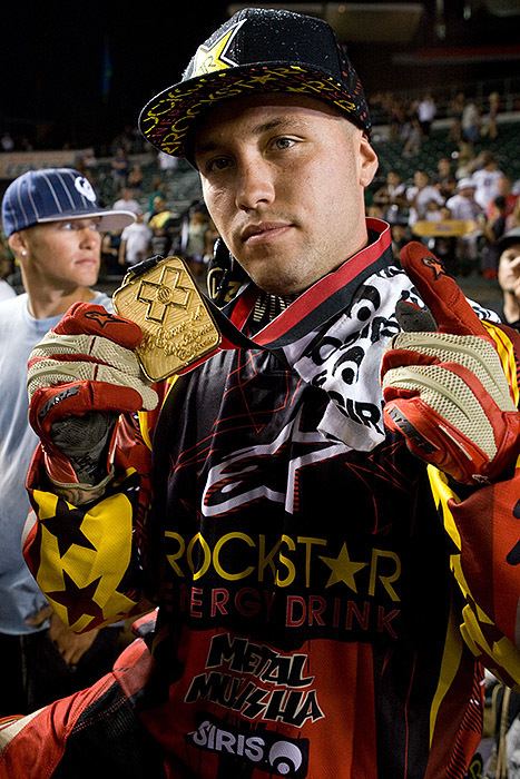 Jeremy in a serious look showing his gold medal with one finger of another hand pointing upwards wearing a yellow & black cap & a white, yellow & black moto racing dress with rockstar written on it.