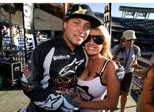 Jeremy and Lauren Lusk both smiling & holding hands at the 2008 ESPN Moto X World Championships in San Diego, California, with a woman in the Background where Jeremy is wearing a cap & moto racing dress, whereas Lauren has wavy hair, wearing big sunglasses, a necklace & a low cut black & white sleeveless top.