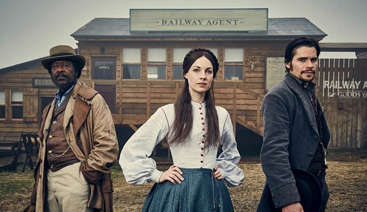 Jessica Raine posing with Clarke Peters and Hans Matheson while behind them is the building of railway agent in the 2016 period drama series Jericho