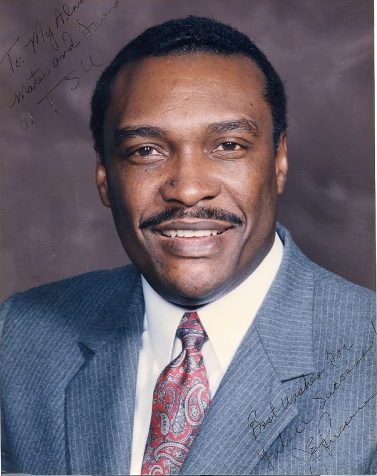 Jesse Russell smiling with a mustache and words written on the top left corner "To: my Alma Mater and Friend at TSU" and "Best Wishes for Future Success with a signature" on the bottom right corner and Jesse wearing a white shirt under a gray coat and a red necktie