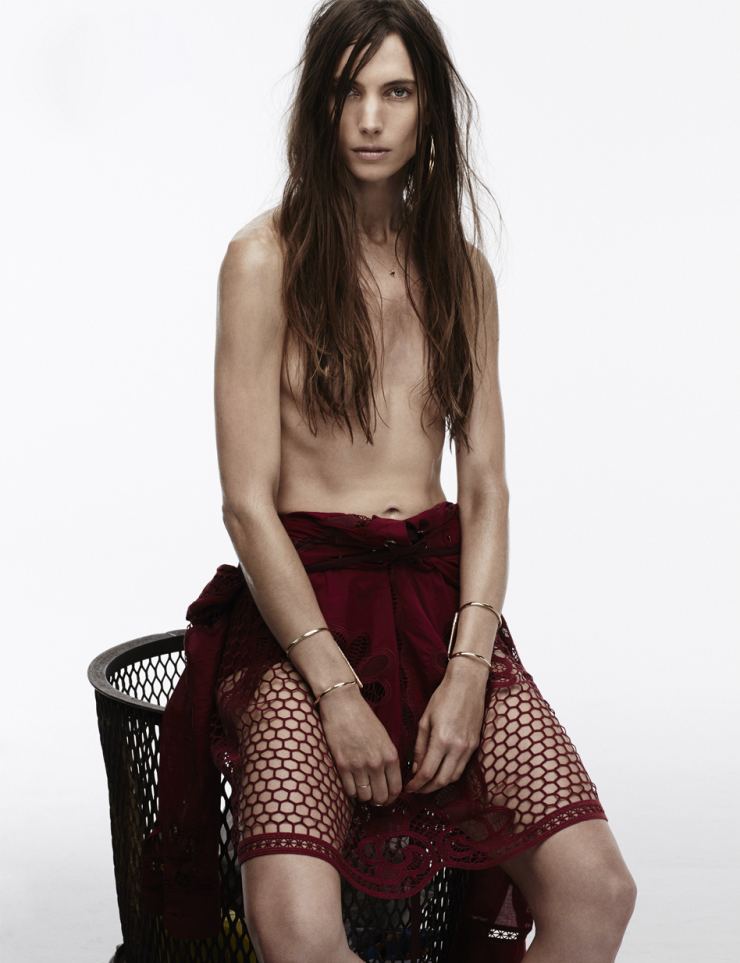 Jessica Miller is topless while sitting on a laundry basket, with a serious face, with long messy hair covering her breasts, and wearing a maroon net skirt.