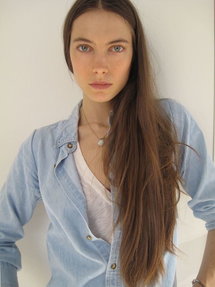 Jessica Miller with a serious face while leaning on the wall, with long hair, wearing a necklace, a denim long sleeves over a white shirt.