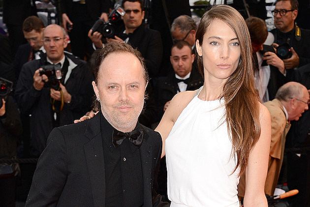 Jessica Miller with a serious face while Lars Ulrich smiling and surrounded by cameramen. Jessica with long hair and wearing a white sleeveless dress while Lars is wearing a black coat over black long sleeves with a black bow tie.