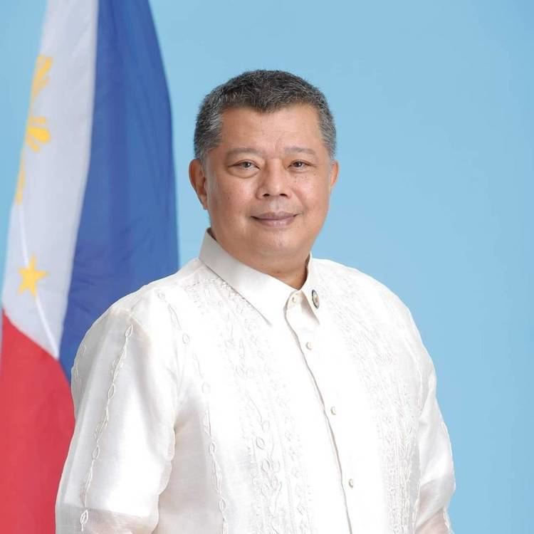 In front of a light blue backdrop with a philippines flag at the left, Jesus Crispin Remulla is smiling, standing, he has black hair wearing a white shirt under a barong tagalog.