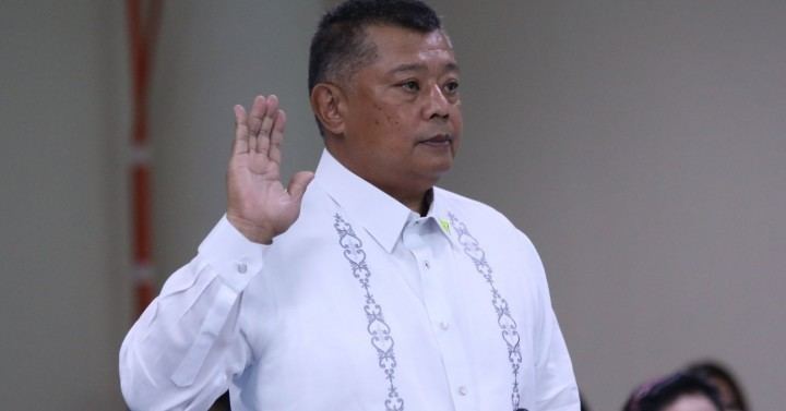 In a room with white walls where Jesus Crispin Remulla is serious, standing, with his right hand up  and swearing solemnly, he has black hair wearing a white barong tagalog.