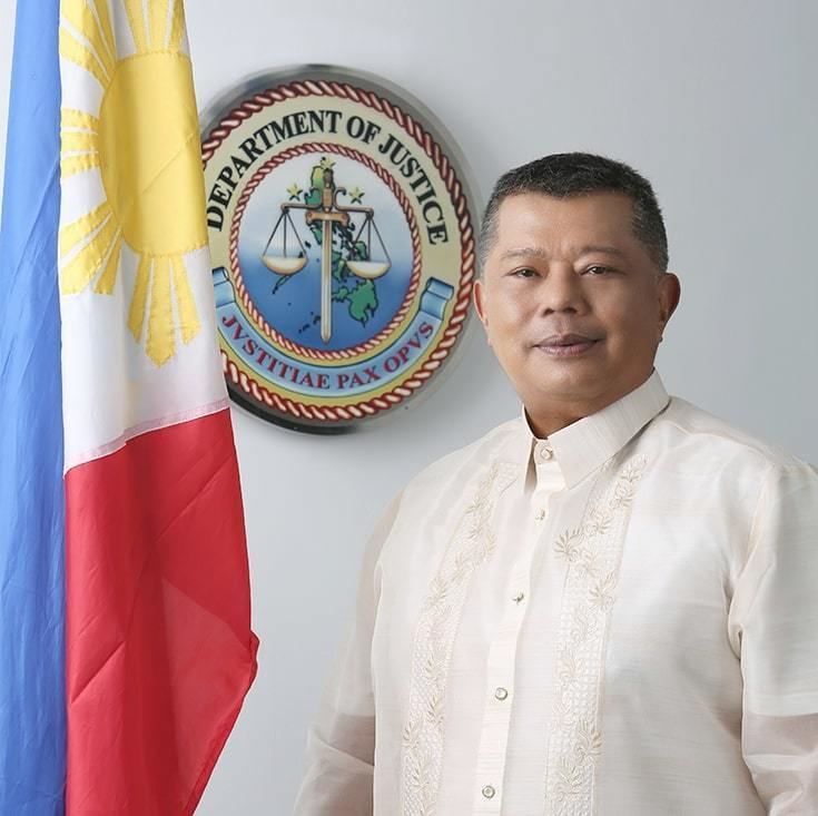 In a white room with a department of justice sign on the wall with a philippines flag at the left, Jesus Crispin Remulla is smiling, standing, he has black hair wearing a white shirt under a barong tagalog.