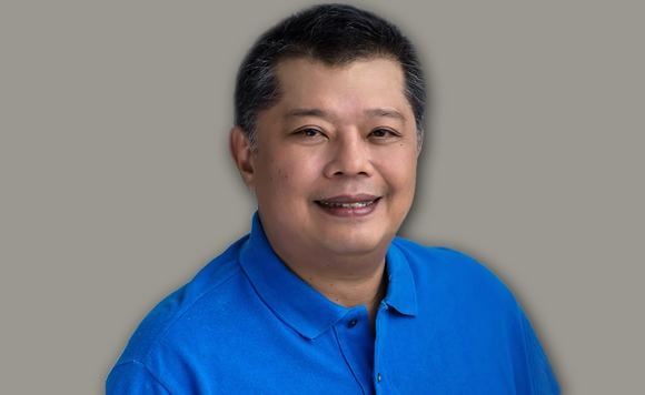 On a gray background, Jesus Crispin Remulla is smiling, standing, he has black hair wearing a blue polo shirt.