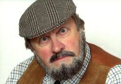 Jethro with mustache and beard while wearing a checkered hat, brown vest and checkered long sleeves