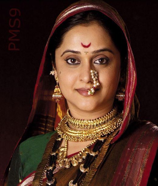 Mrinal Kulkarni as Rajmata Jijabai in the 2008 Marathi historical TV drama, Raja Shivchatrapati, smiling with a bindi on her forehead while wearing a maroon and green saree, pearl nath, gold necklaces, and earrings