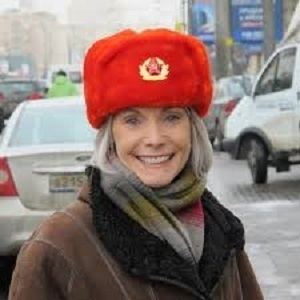 Jill Dougherty smiling on a street and wearing a black and brown coat with a scarf and orange bonnet hat