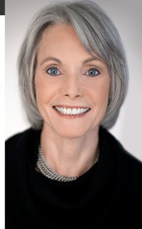Jill Dougherty smiling with white short hair and wearing a black coat and bead necklace