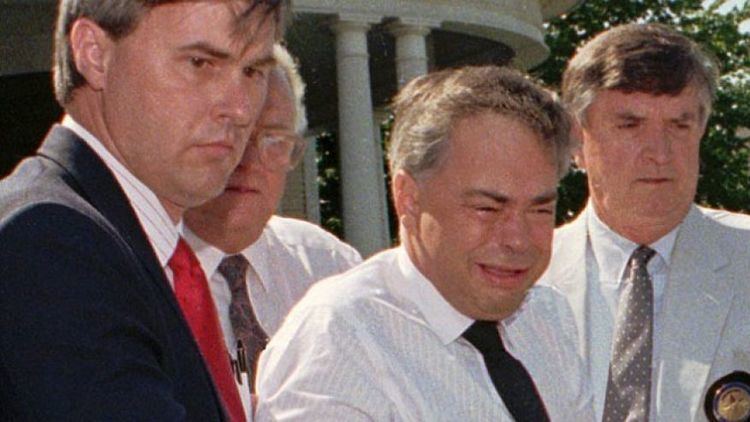 Jim Bakker crying while being hauled off to jail by several men and wearing a white shirt and a black necktie