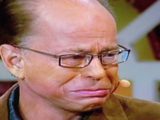 Jim Bakker looking distressed while wearing eyeglasses and a blue shirt under a brown coat