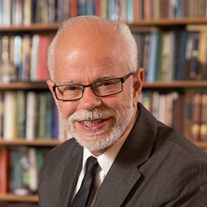 Jim Bakker smiling with a mustache and a beard while wearing eyeglasses, white sleeve, gray suit and a black necktie