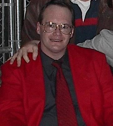 Jim Cornette smiling with a hand on his shoulder, wearing eyeglasses, a red suit, black long sleeves, and a red tie.