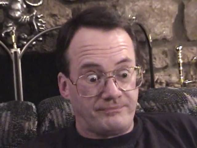 Jim Cornette wearing a surprised-looking expression while sitting on a couch, wearing eyeglasses, and a black shirt.
