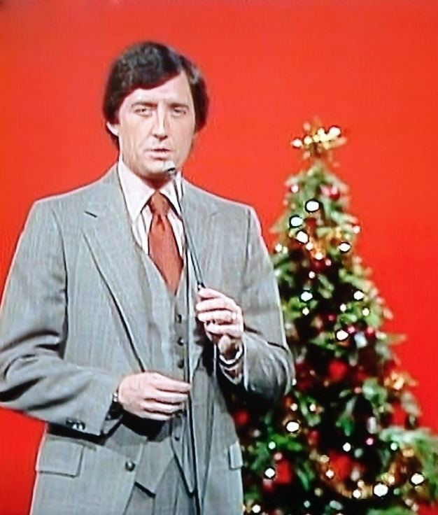Jim Perry holding a microphone while wearing a gray coat and red necktie in an orange background