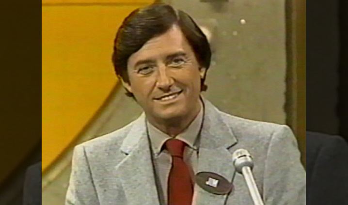 Jim Perry smiles with a microphone in front of him wearing a gray coat and a red necktie