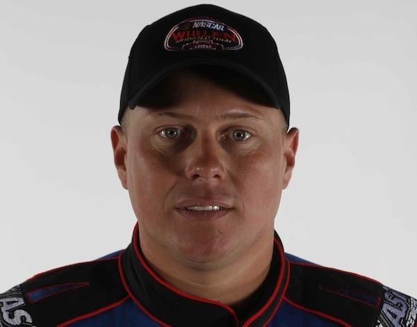 In a white background, Jimmy Blewett is serious, standing, wearing a black race cap with nascar broided on it, and a black race tracksuit.