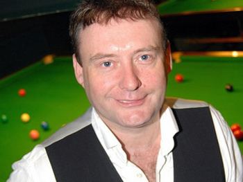 Jimmy White smiling in front of a pool table with balls on it and wearing a black vest over white long sleeves.