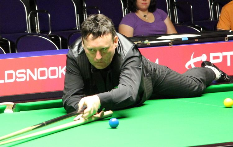 Jimmy White with a serious face while lying on the pool table with balls on it and holding cue sticks, wearing a black vest over black long sleeves, black bowtie, black pants, and black shoes.