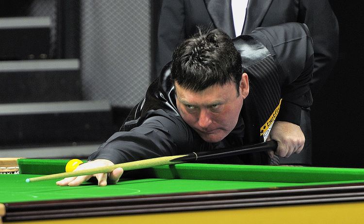 Jimmy White with a serious face while playing pool and holding a cue stick and wearing a black vest over black long sleeves.