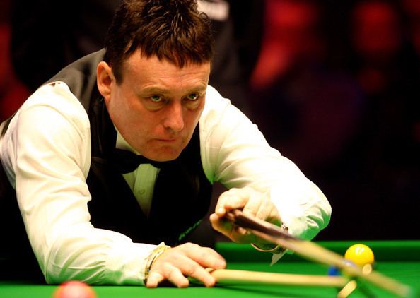 Jimmy White with a serious face while leaning on a pool table with balls on it and holding cue sticks, wearing a black vest over white long sleeves and black bowtie.