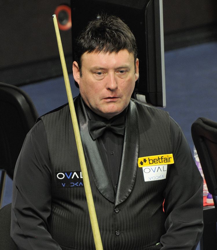 Jimmy White with a serious face while looking at something, with a cue stick on his shoulder, wearing a black vest over black long sleeves and black bowtie.
