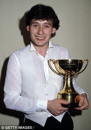 Jimmy White is smiling while holding a trophy in the Masters' Snooker Championship Final at Wembley Arena in London on 27th January 1984. Jimmy is wearing a white vest over white long sleeves and black pants.