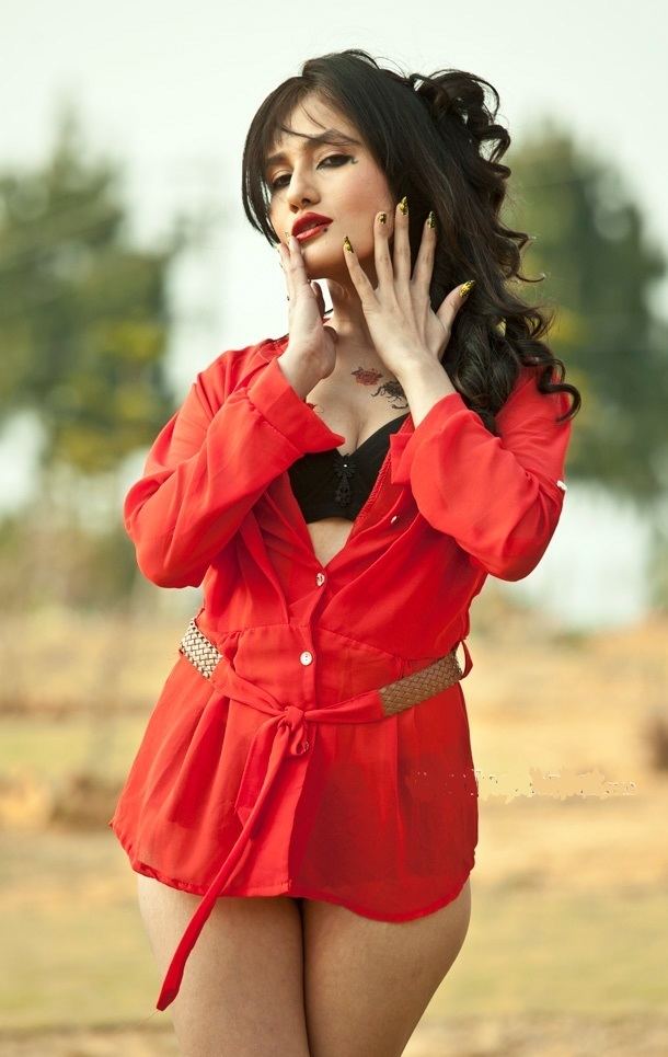 Jiya KC is serious, standing, both hands up open under her jaws, has long black curly hair, black fingernails, tattoo on her left chest, wearing a red showing cleavage long sleeve dress, and a black bra.