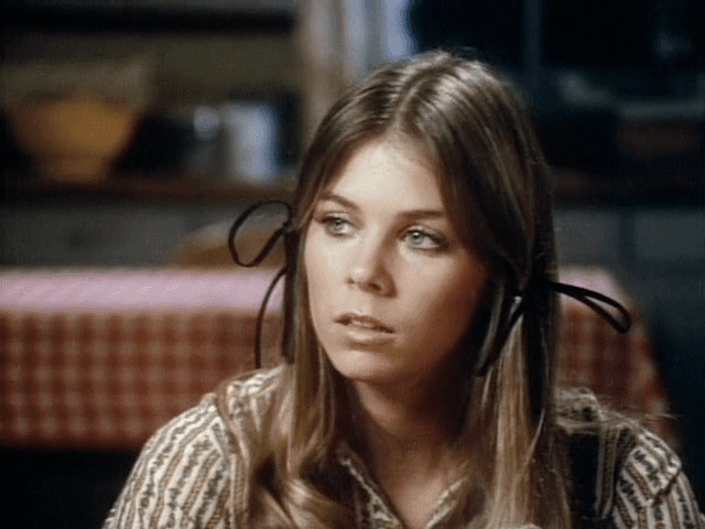 Jo Ann Harris looking at something and having ribbons in her hair and wearing a striped, floral designed dress.