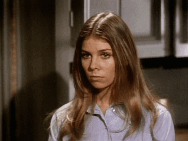 Jo Ann Harris looking at something inside a house and wearing a sky blue buttoned dress.