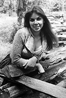 Jo Ann Harris smiling while posing in some wood and wearing a long sleeveless dress.