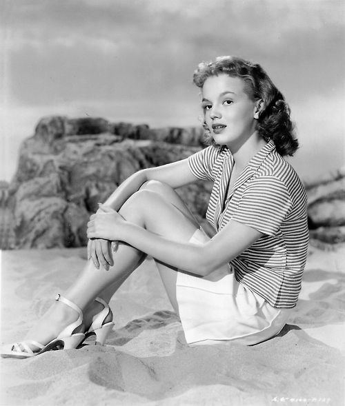 Joan Evans looking afar while sitting on the sand and wearing a striped blouse, skirt, and sandals